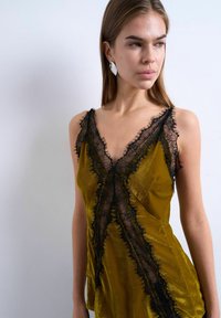 Jeune femme aux longs cheveux bruns portant un caraco en satin moutarde avec des bordures en dentelle noire, regardant sur le côté contre un fond clair uni.