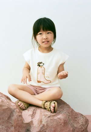 Jeune fille assise en tailleur sur un rocher, portant un débardeur blanc avec un motif de botte et un short rose.