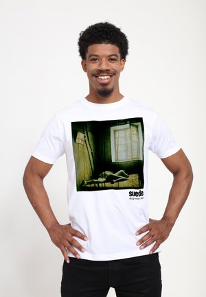 Man smiling with hands on hips wearing white t-shirt showing art of a figure lying by a window with "suede dog man star" text.
