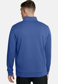 Uomo che indossa una giacca blu con zip a maniche lunghe, di spalle, su uno sfondo bianco semplice.