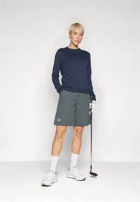 Marinblå långärmad tröja i kombination med grå knälånga shorts med en logotyp. Vita sportskor och en golfklubba ingår.