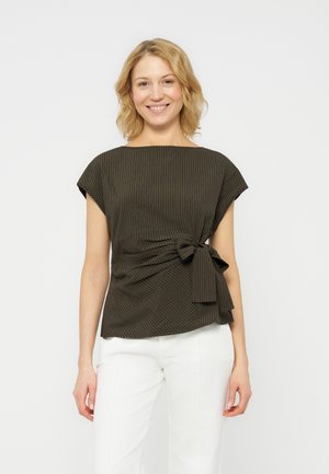 Usmievavá žena s blond vlasmi po plecia, ktorá má na sebe tmavozelený vrchný diel s tenkými pásikmi a bočnou mašľou, a biele nohavice, stojaca na bielom pozadí.