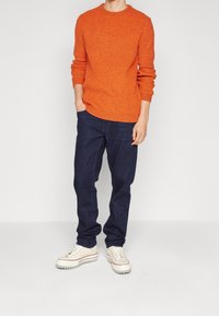 Maglione arancione lavorato a maglia con scollo rotondo, abbinato a jeans di denim blu scuro. Le scarpe sono delle sneakers crema con suola spessa e dettagli rossi.