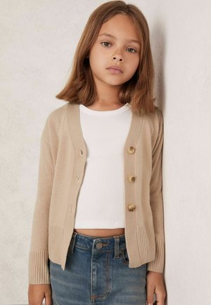 Beige Strickjacke mit vier großen Knöpfen, gerippten Bündchen und Rundhalsausschnitt, getragen über einem weißem enganliegendem Oberteil und Jeansshorts.