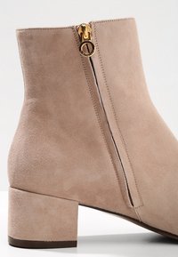 Botín de ante en color beige claro, con una cremallera lateral con un acento dorado y un tacón grueso. Textura suave con mínimos detalles de costura.