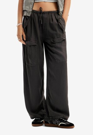 Persona con pantalones cargo negros holgados con cintura ajustable con cordón, bolsillos laterales y solapas en el muslo, combinados con zapatillas negras y top corto.