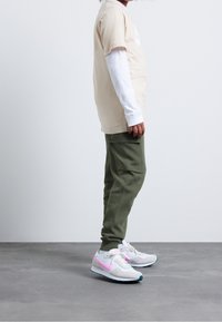 Ett barn i beige T-shirt över en vit långärmad tröja, gröna cargo-joggers och vita sneakers med rosa detaljer, står på en grå golv.
