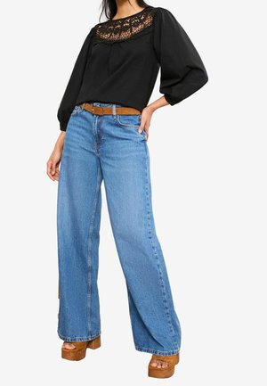 Femme portant un chemisier noir avec empiècement en dentelle, jean bleu à jambes larges, ceinture marron et sandales compensées marron, debout avec une main sur la hanche.