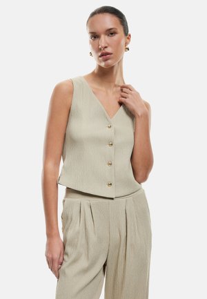 Beige ilmanhihaisessa topissa on V-pääntie, viisi kultanappia, teksturoitu kangas ja lyhyt malli. Yhdistetty pleitettyihin beigeihin housuihin.