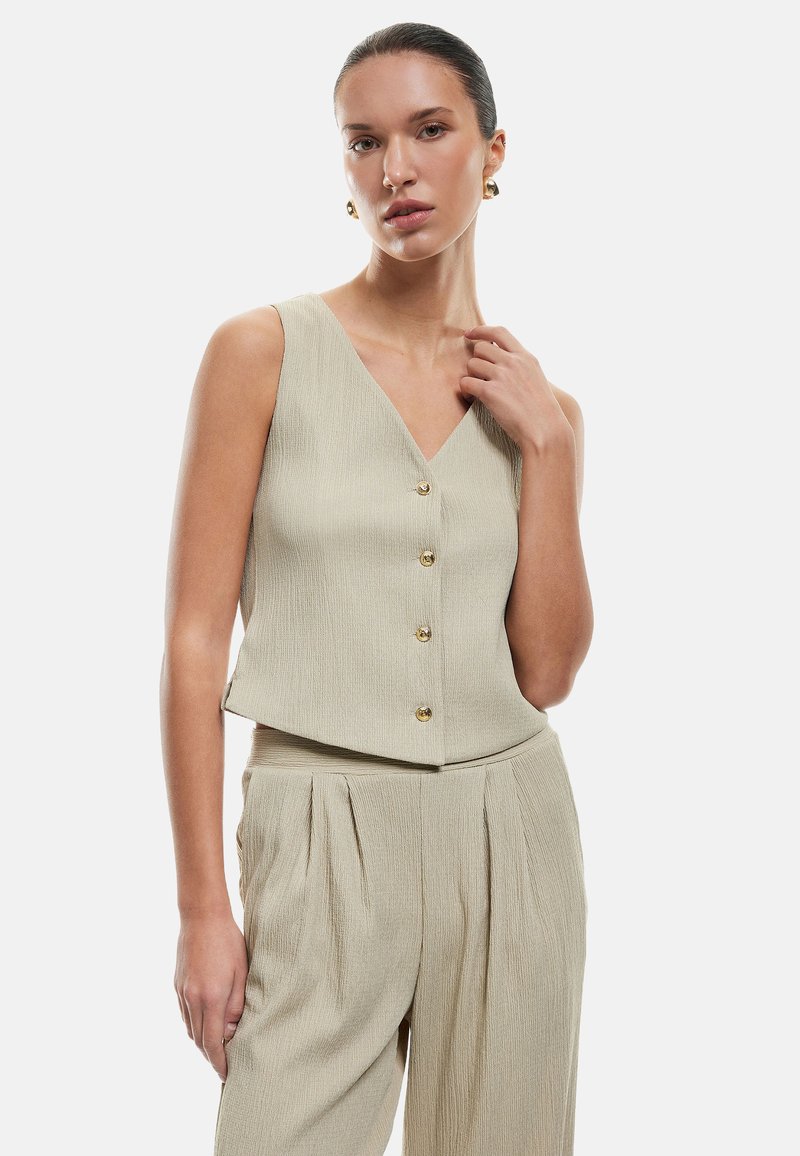 Beige ilmanhihaisessa topissa on V-pääntie, viisi kultanappia, teksturoitu kangas ja lyhyt malli. Yhdistetty pleitettyihin beigeihin housuihin.