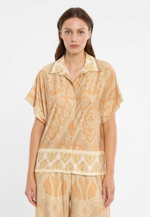 Femme portant une chemise beige ample à manches courtes avec des boutons, ornée d'un motif cachemire orange, et un pantalon assorti, posant devant un fond blanc.