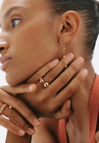 Las joyas de oro incluyen un pendiente colgante con acentos esféricos y dos anillos, uno con una esfera pulida. La piel tiene una textura natural y suave.