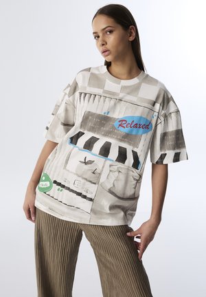 T-shirt grafico a maniche corte in tonalità chiare presenta vari estampati, tra cui il testo "Rilassato" e "Gusto", abbinata a pantaloni olive testurizzati e plissettati.