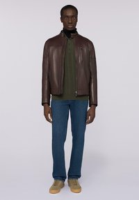 Giacca di pelle marrone con colletto alto, chiusura con zip, indossata sopra un maglione a coste verde scuro, abbinata a jeans blu e sneakers chiare.