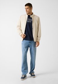 Beige Bomberjacke mit Reißverschluss über einem marineblauen Pullover, kombiniert mit hellblauen Jeans und marineblauen Sneakern mit beigen Akzenten. Glatter Stoff, lässiges Design.