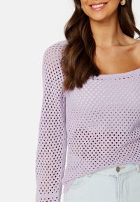 Pullover di maglia viola chiaro con design a rete aperta, maniche lunghe e scollo ampio, abbinato a pantaloni di denim blu chiaro.
