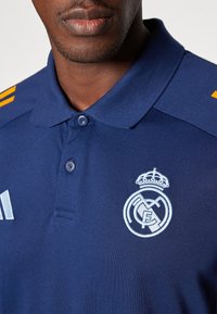 Gros plan sur une personne portant un polo bleu avec un logo Real Madrid blanc et des rayures jaunes sur l'épaule.