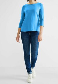 Sininen, pitkähihainen pusero pehmeästä ja sileästä kankaasta, jossa on leveä kaula-aukko. Yhdistetty tummaansinisiin farkkuihin ja valkoisiin lenkkareihin, luoden rentoa ilmettä.