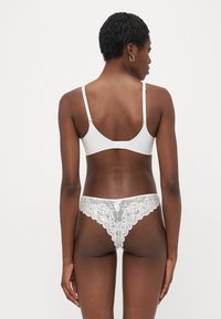 Vrouw die van achteren staat, in een witte beha met verstelbare bandjes en een witte kanten slipje met bloemenmotief, tegen een effen achtergrond.