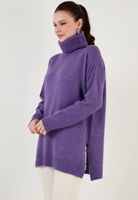 Lila pulover s turtleneck ohlapnega kroja, z rebrastimi manšetami in robom, stranskimi razporki ter mehkimi tkaninami. Oblečen z svetlobe obleko.