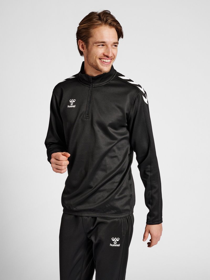 Giovane uomo sorridente, indossa una tuta sportiva nera Hummel con chevron bianchi sulle spalle, su uno sfondo grigio uniforme.
