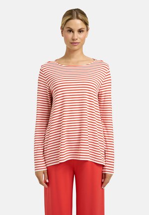 Frau trägt ein weißes Langarmshirt mit dünnen roten horizontalen Streifen und rote, weit geschnittene Hosen, steht vor einem einfarbigen Hintergrund.