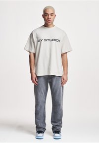 Light beige oversized t-shirt with black "2Y STUDIOS" text, paired with gray denim jeans and blue and black sneakers.