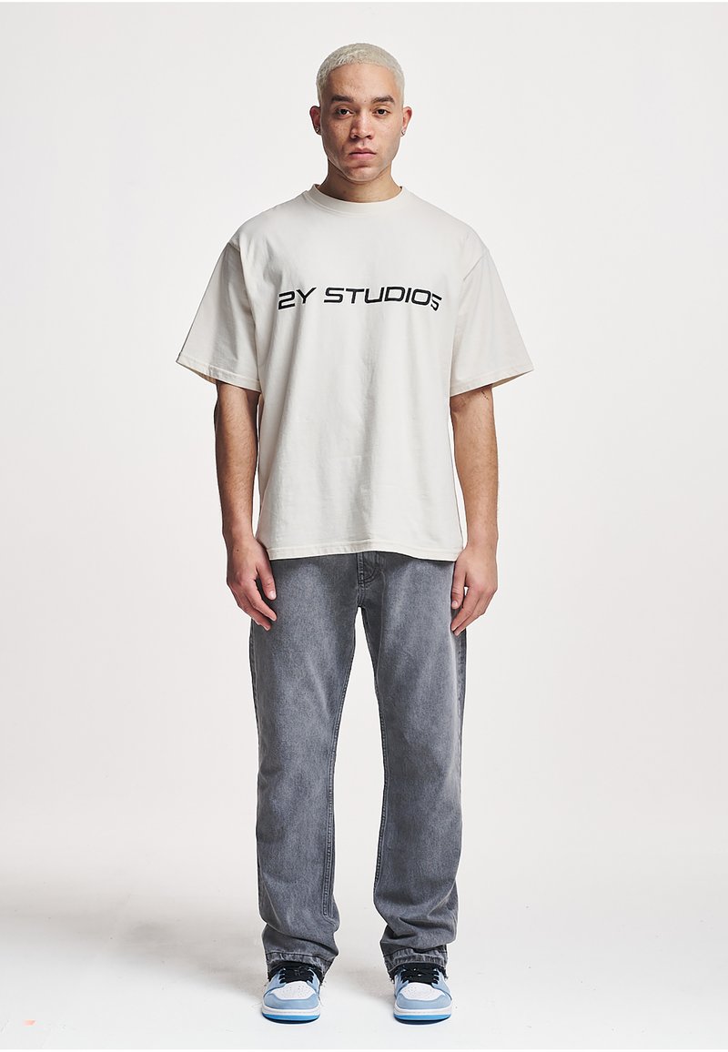 Light beige oversized t-shirt with black "2Y STUDIOS" text, paired with gray denim jeans and blue and black sneakers.