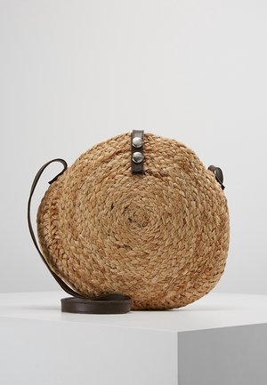 Okrągła pleciona słomiana torba na ramię z ciemnobrązowym skórzanym paskiem i zapięciem na zatrzask, na jednolitej białej powierzchni.