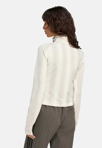 Biela blúzka s dlhými rukávmi, vertikálnymi pruhmi, vysokým golierom a priliehavým strihom, doladená s tmavými pruhovanými nohavicami. Hladká textúra a tesný strih.