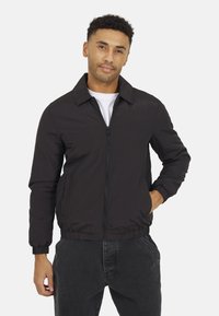 Giacca bomber leggera nera con colletto, chiusura zip, polsini elasticizzati e tasche laterali, indossata sopra una maglietta bianca. Texture liscia.