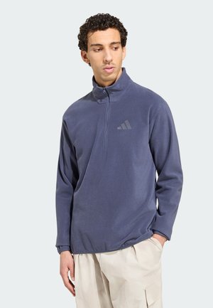 Uomo con capelli ricci che indossa un pile blu navy con zip al quarto e pantaloni beige, guarda verso sinistra con una mano in tasca.