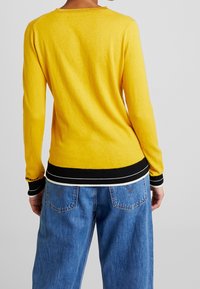 Geel gebreid sweater met een ronde hals en een geribbelde zwarte zoom. Heeft lange mouwen met zwart-wit accenten bij de manchetten. Gedragen met blauwe jeans.
