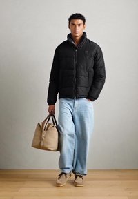 Chaqueta de plumas negra con cierre de cremallera y costuras horizontales, combinada con jeans baggy de color azul claro y zapatos beige; sosteniendo un bolso beige.