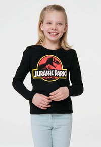 Schwarzes Langarm-T-Shirt mit einem bunten "Jurassic Park"-Grafikdesign und einem Dinosaurier-Emblem. Kombiniert mit hellblauen Jeans.