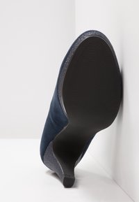 Escarpin en daim bleu marine avec un accent argenté scintillant au talon, doté d'une semelle en caoutchouc noire texturée. Forme élégante et effilée.
