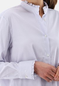 Camicia a righe viola chiaro e bianche con colletto volant, chiusura a bottoni e polsini con bottone. Presenta una vestibilità rilassata e una texture del tessuto liscia.