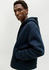 Marinblå zip-up hoodie i mjukt tyg, med rymlig huva, sidofickor och ribbade muddarna och fållen. Mjuk textur och avslappnad passform.