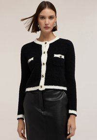 Svart stickad cardigan med vit kant, försedd med guldknappar och två bröstfickor. Strukturerad tyg, kort design och långa ärmar.