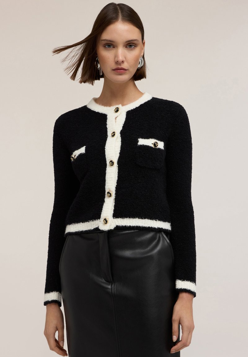 Svart stickad cardigan med vit kant, försedd med guldknappar och två bröstfickor. Strukturerad tyg, kort design och långa ärmar.