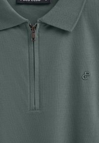 Mörkgrön pikétröja med en dragkedjekrage och broderad logotyp på vänster sida. Gjort av mjukt, texturerat tyg.