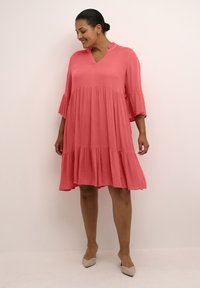 Mujer vestida con un vestido de coral rosa hasta la rodilla, con mangas acampanadas y tacones beige de punta, de pie contra una pared clara.