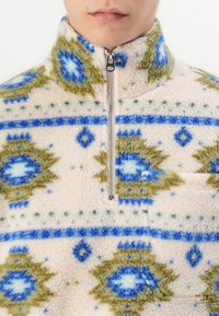 Korkeakauluksinen fleece-takki, jossa on puoli-vetoketju ja sinivihreitä geometrisia kuvioita vaalean beigen taustan päällä.