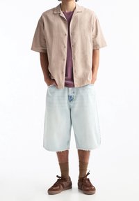 Hombre con una camisa de botón de manga corta beige sobre una camiseta morada, pantalones cortos de mezclilla azul claro hasta la rodilla, calcetas marrones y zapatillas marrones.