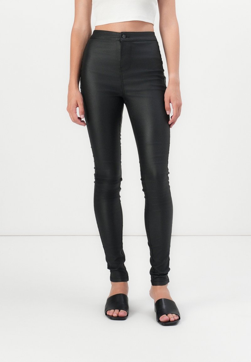 Femme portant un pantalon skinny en cuir noir taille haute et des sandales ouvertes noires, debout devant un fond blanc.
