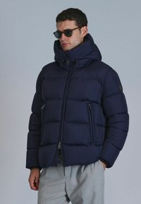 Marineblaue Steppjacke mit hohem Kragen, Reißverschlusstaschen und strukturiertem Quiltmaterial. Schmaler Schnitt, kombiniert mit hellgrauen Hosen. Heller Hintergrund.