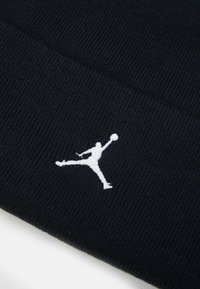 Svart strikket lue med ribbet tekstur, med en fremtredende hvit brodert Jumpman-logo. Ingen andre detaljer eller mønstre er til stede.