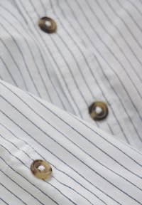 Camicia di cotone bianca con sottili righe blu navy, dotata di bottoni rotondi marroni e una superficie liscia. Bottoni posizionati lungo la patta della camicia.