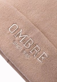 Beige neulekangas, jossa on pehmeä rakenne, ja jossa on brodeerattu teksti "OMBRE" ja "SINCE 1926" vaaleilla langoilla.