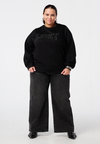 Sweatshirt noir avec "LUCKY" en lettres scintillantes, associé à un jean noir à jambes larges. Caractéristiques : coupe décontractée et tissu texturé.